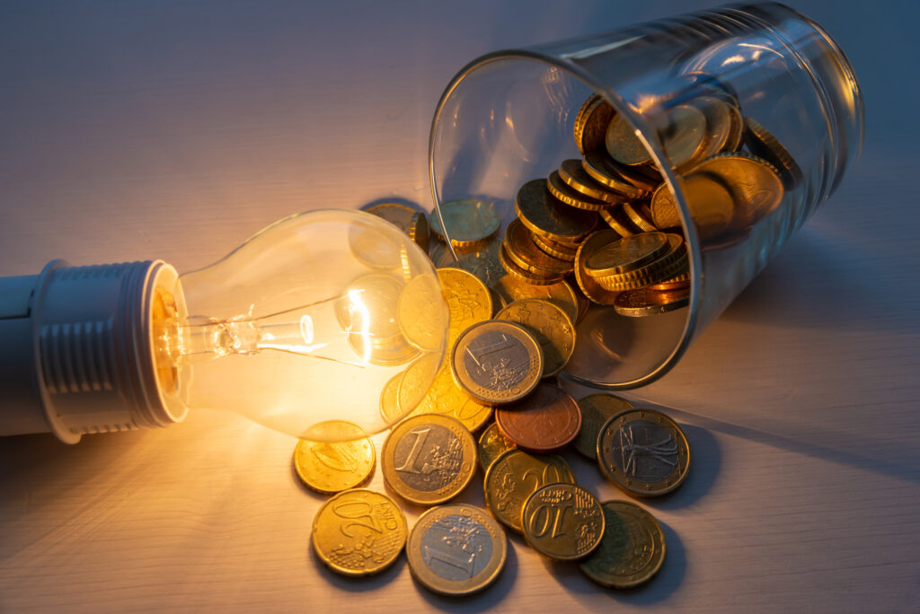 Ein umgekipptes Glas und daraus neben eine Lampe fallende Euromünzen zeigen Stromkosten an.